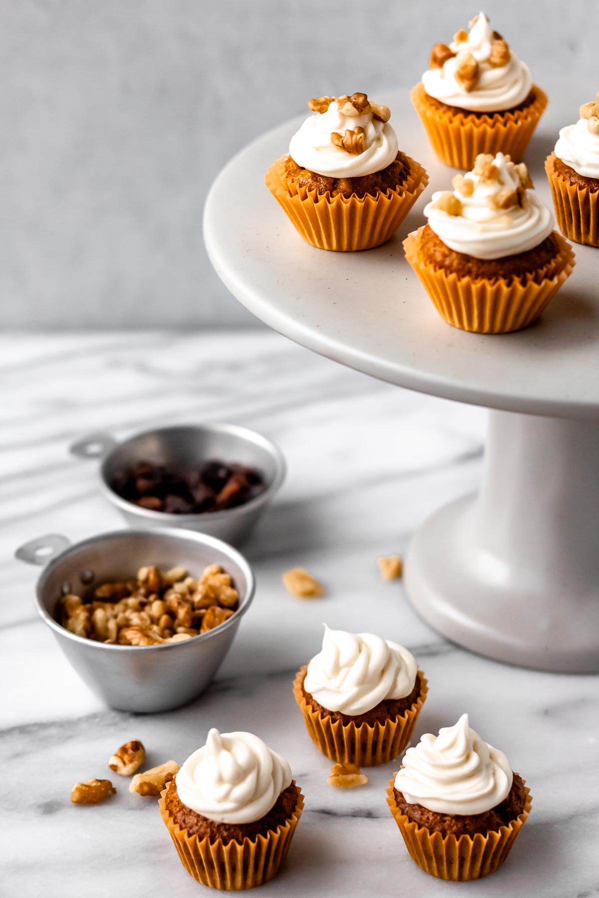 Mini carrot cakes on and around a cake stand with 2 measuring cups filled with raisins and walnuts.