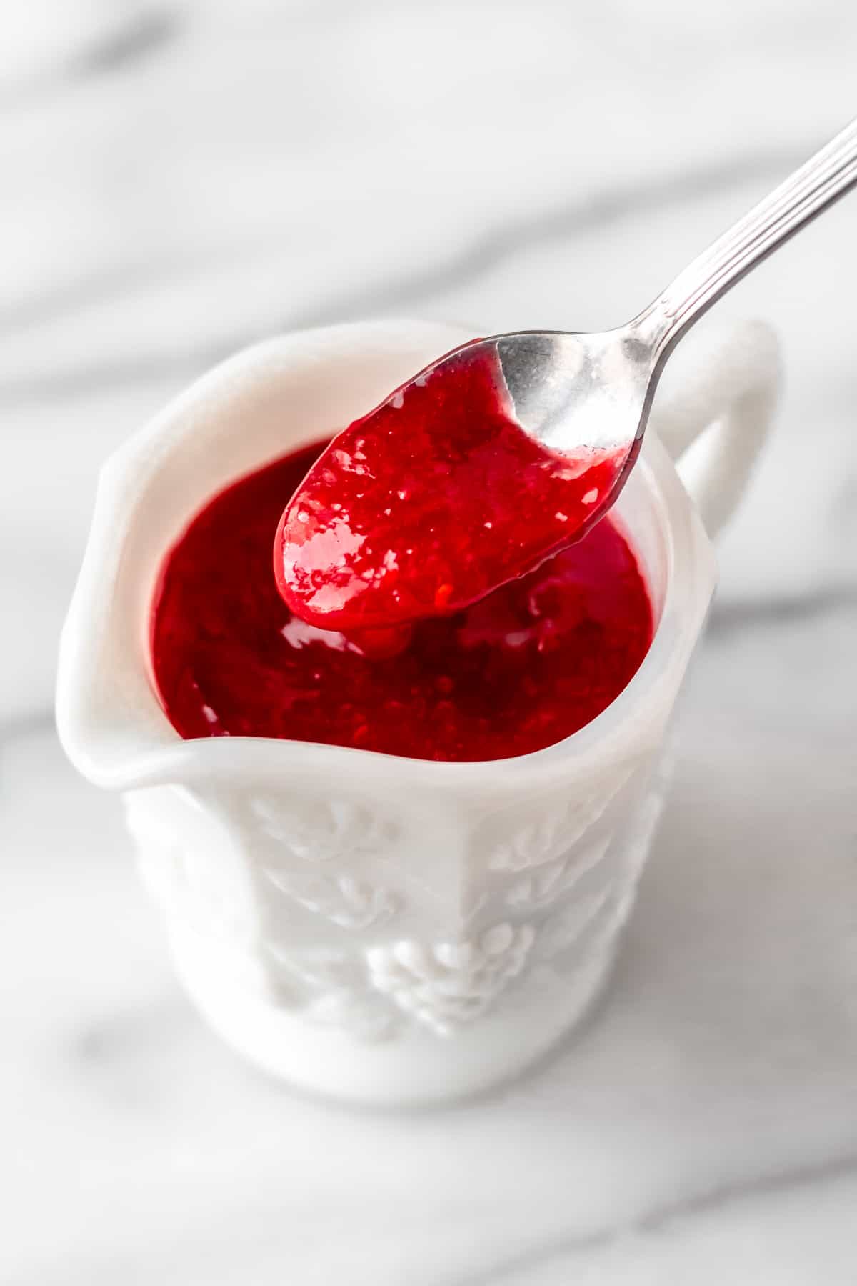 A spoonful of homemade raspberry sauce being lifted up over a cup of sauce.