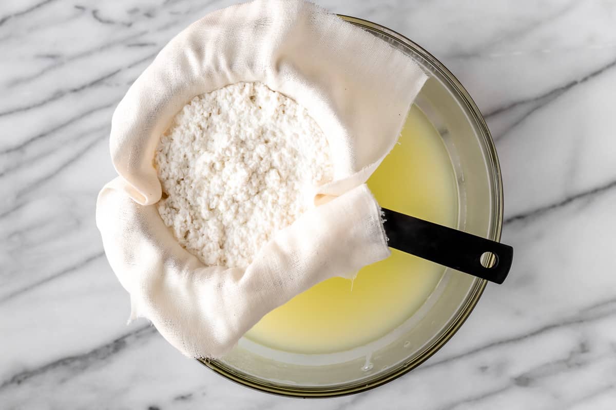 Cheese curds being strained through a cheesecloth lined sieve over a bowl of whey.