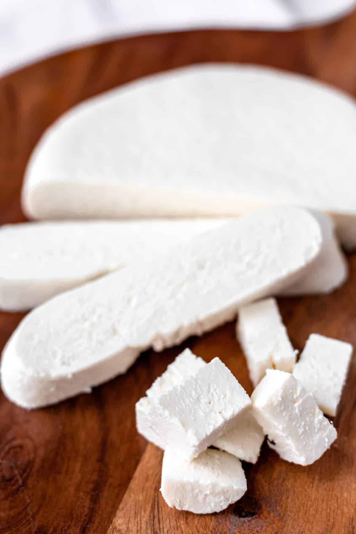 Close up of a chunk of paneer with two slices and many cubes cut from it on a wood cutting board.