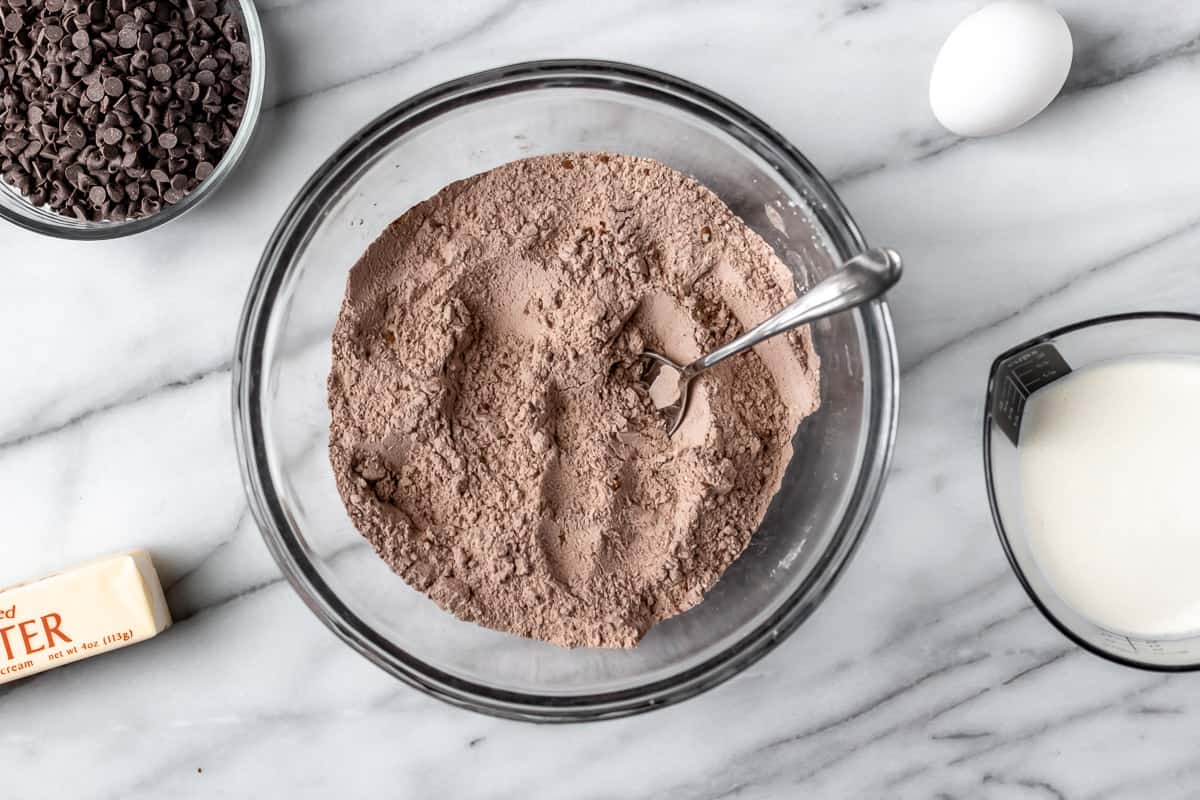 Dry ingredients for chocolate scones in a glass bowl with a spoon and other ingredients around it.