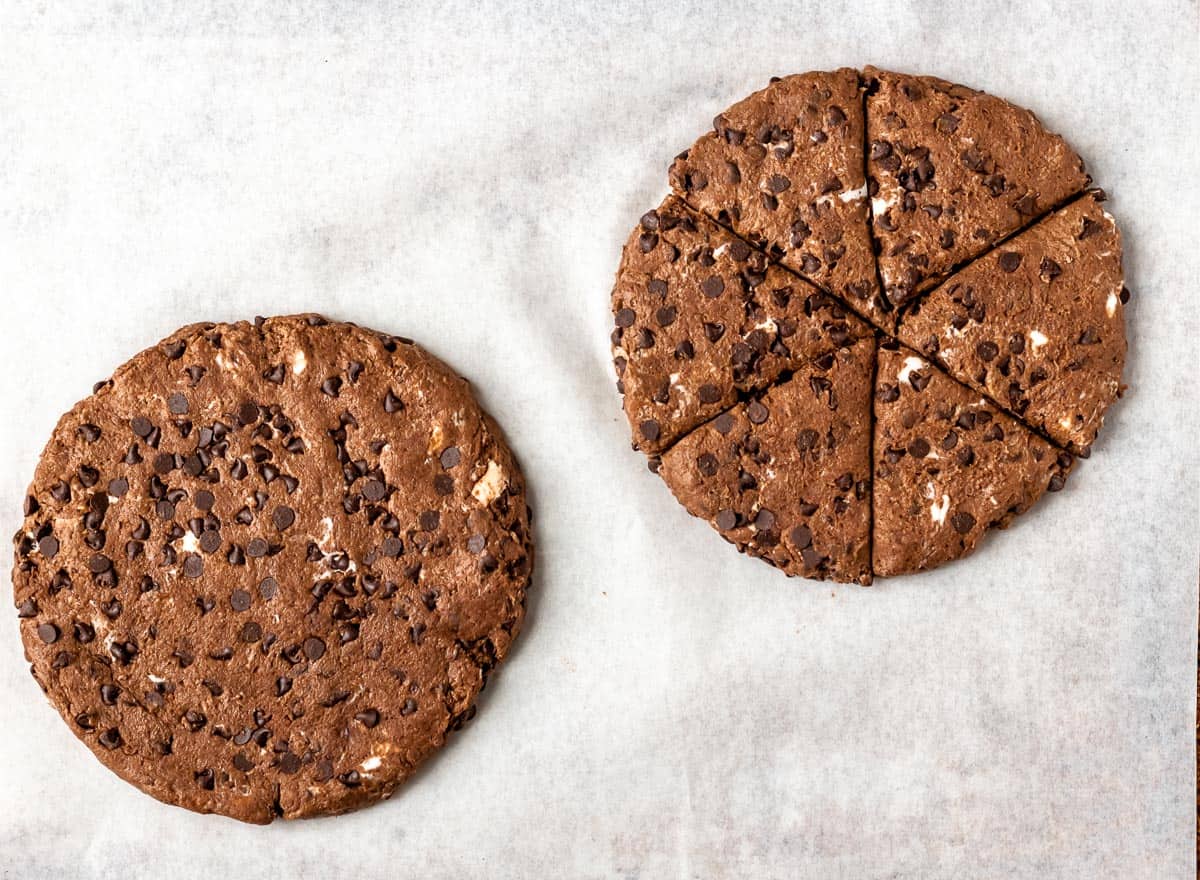 Chocolate scones dough shaped into two discs on a parchment paper with one cut into 6 triangles.