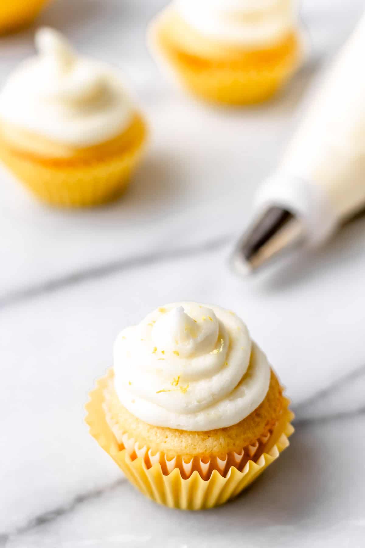 A cupcake with Lemon Buttercream Frosting piped onto it with the piping bag and a few more cupcakes in the background.