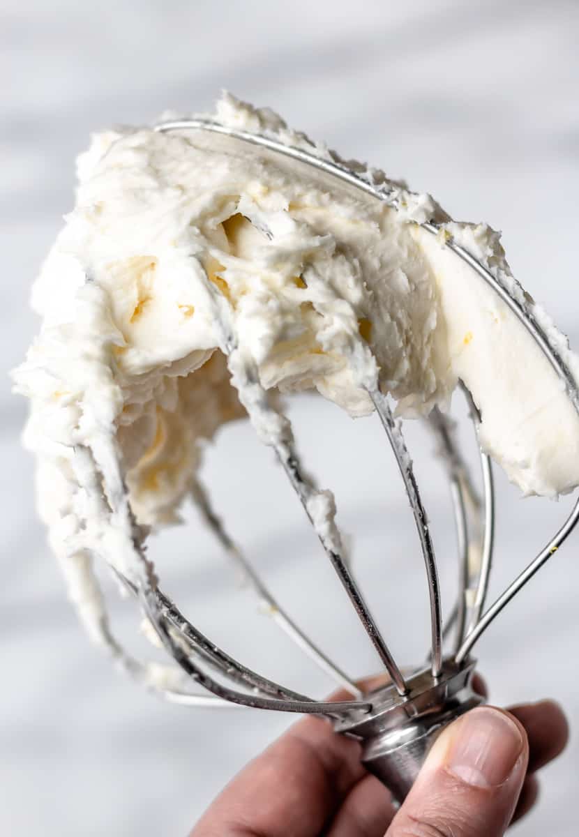 A whisk attachment being held up with Lemon Buttercream Frosting on it.
