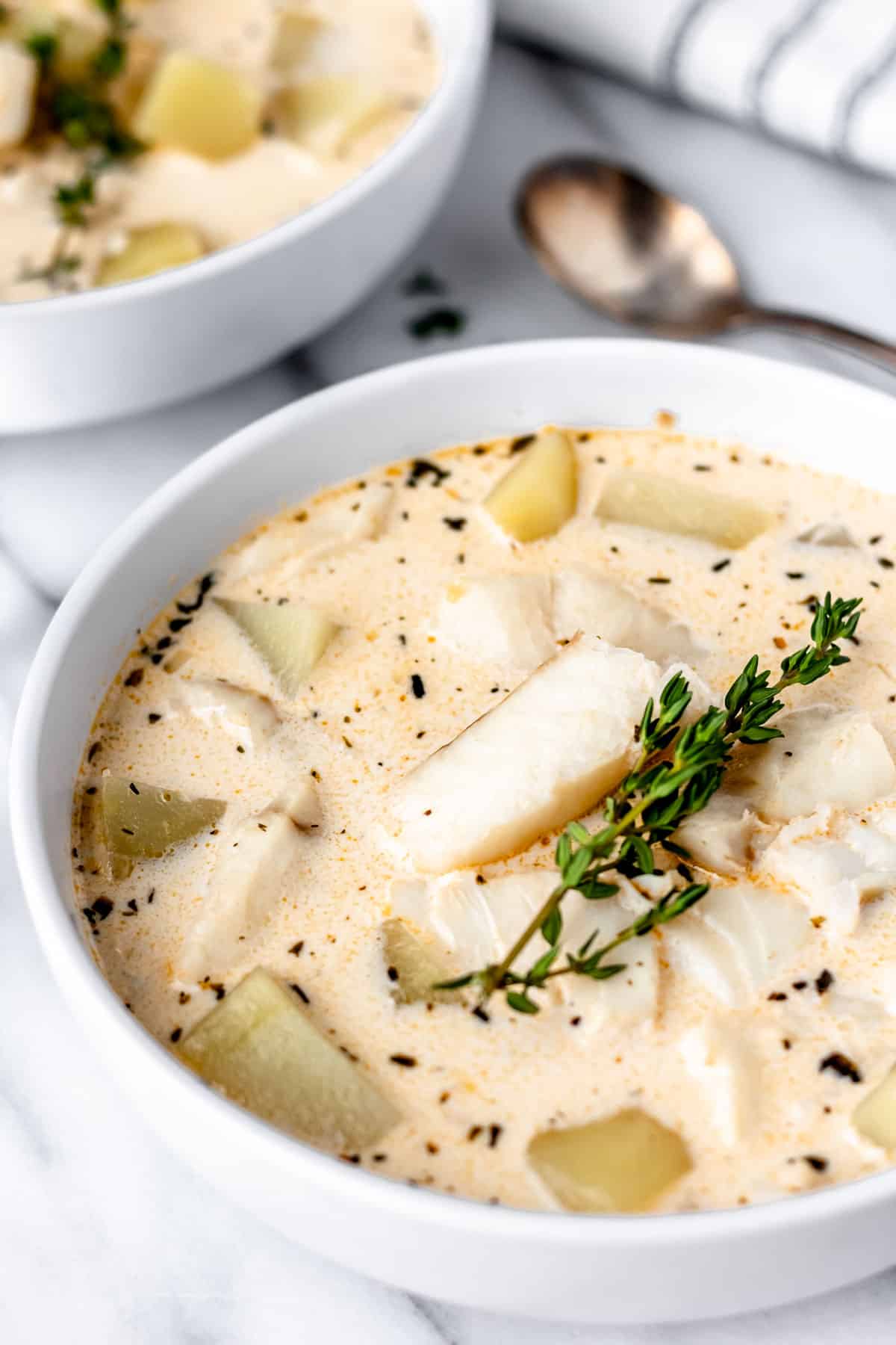 Close up of cod chowder in a white bowl with a second bowl in the background along with a spoon and striped towel.