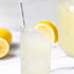 A glass of lemonade over ice with a pitcher of lemonade and lemons in the background.