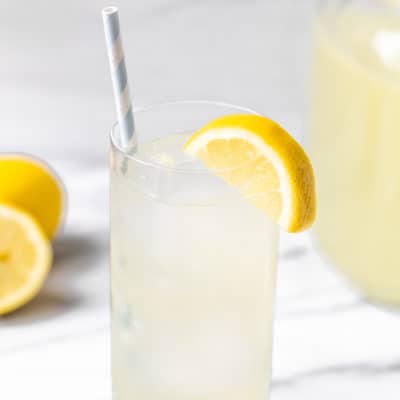 A glass of lemonade over ice with a pitcher of lemonade and lemons in the background.