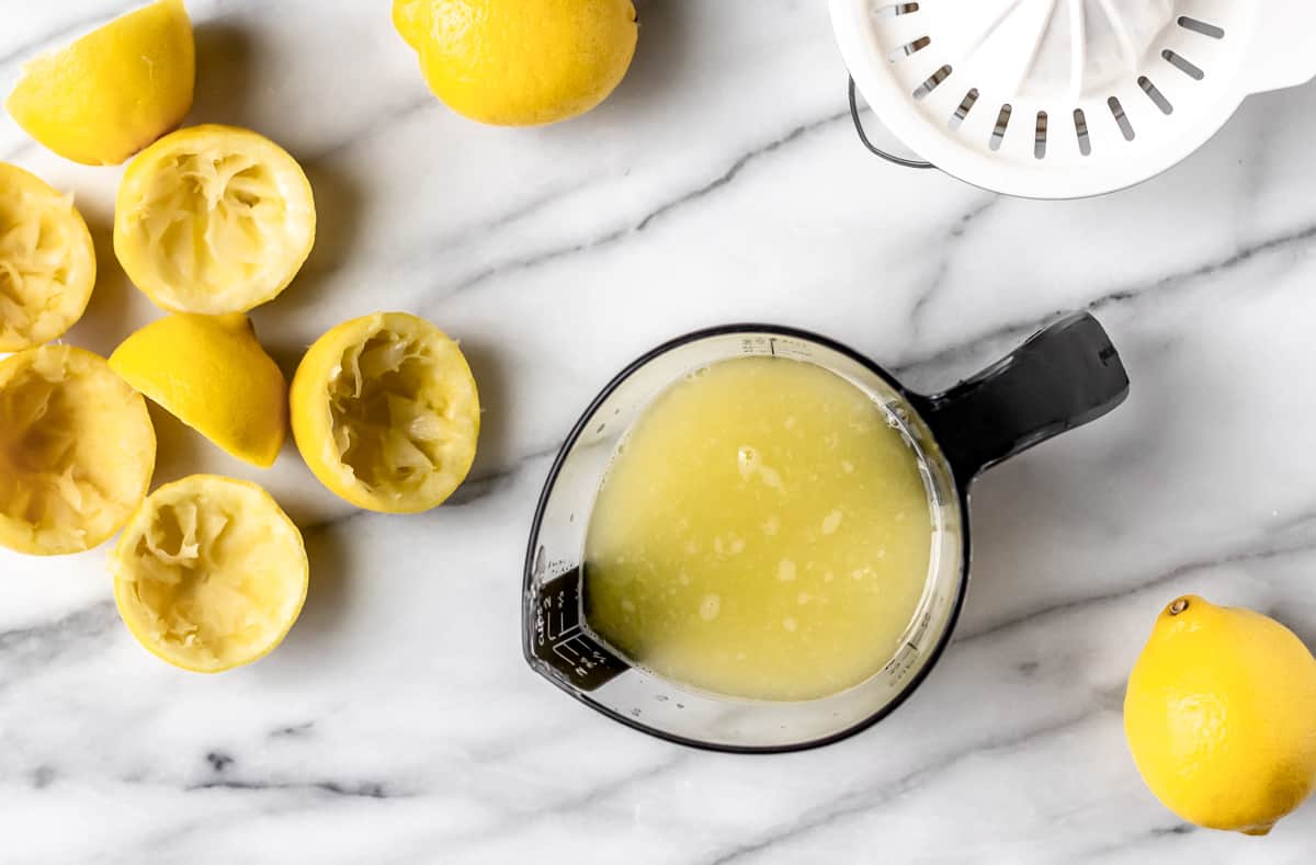 Squeezed lemons with a measuring cup of lemon juice, whole lemons and a juicer on a marble background.