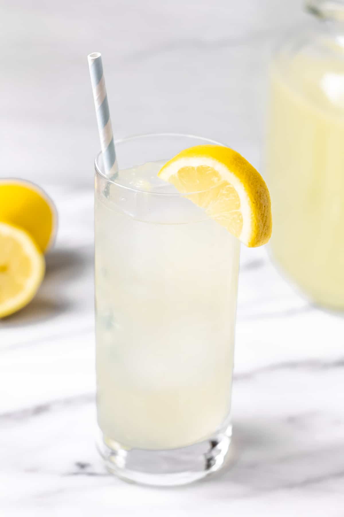 A glass of lemonade with ice in it and a lemon slice on the rim. There is part of a pitcher of lemonade and lemons in the background. 