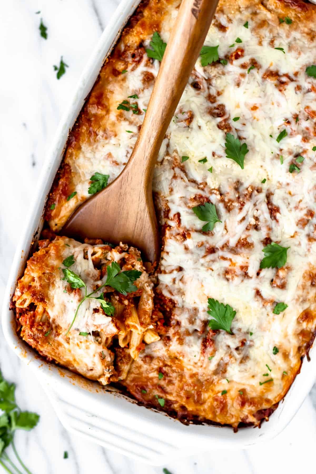 Overhead of a casserole dish of baked ziti with a server lifting up a serving.