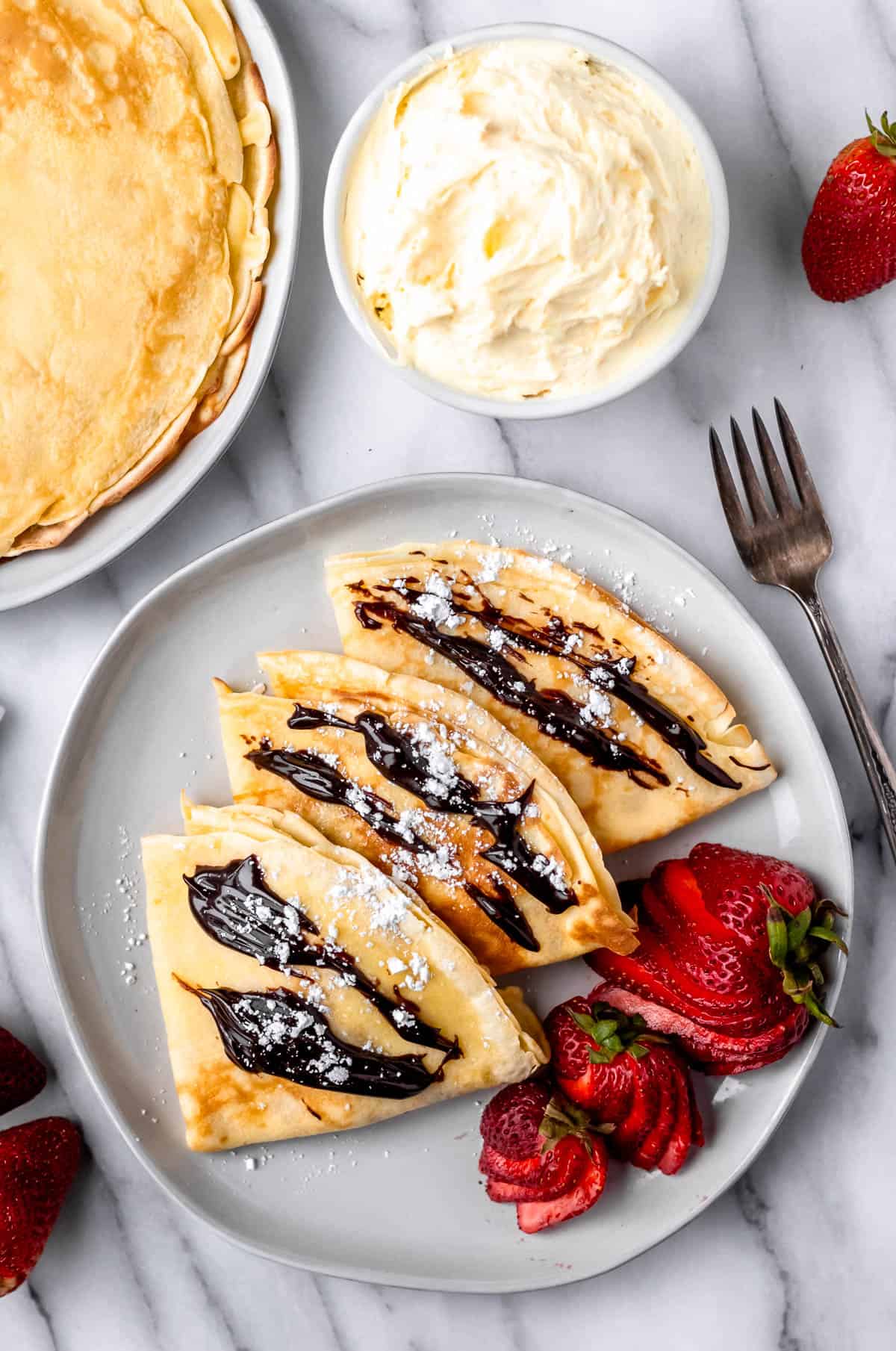 Overhead of a plate of sweet crepes with strawberries, a bowl of pastry cream and a plate of plain crepes around it.