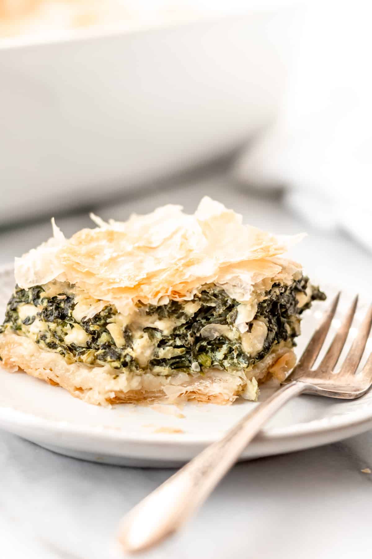 A serving of spanakopita on a white plate with a fork and a white casserole dish partially showing in the background.