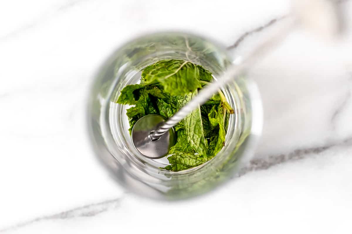 Mint leaves in the bottom of a glass with a metal muddler inside as well.