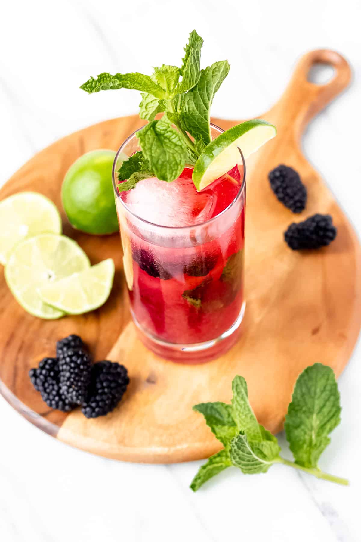 A blackberry mojito on a circular wood board with limes, blackberries and mint leaves around it.