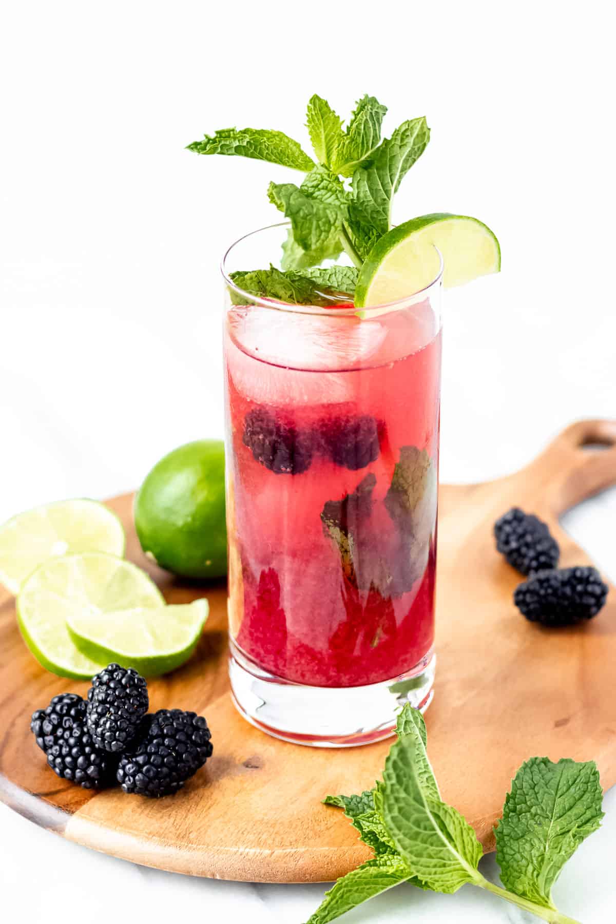 A blackberry mojito on a circular wood board with limes, blackberries and mint leaves around it.