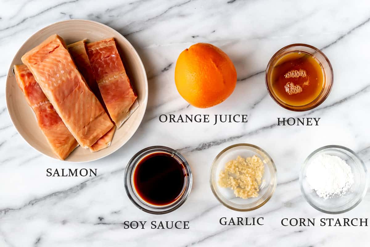 Ingredients to make orange glazed salmon on a marble background with text overlay.
