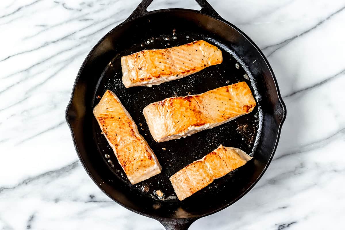 Four salmon filets cooking in a cast iron skillet.