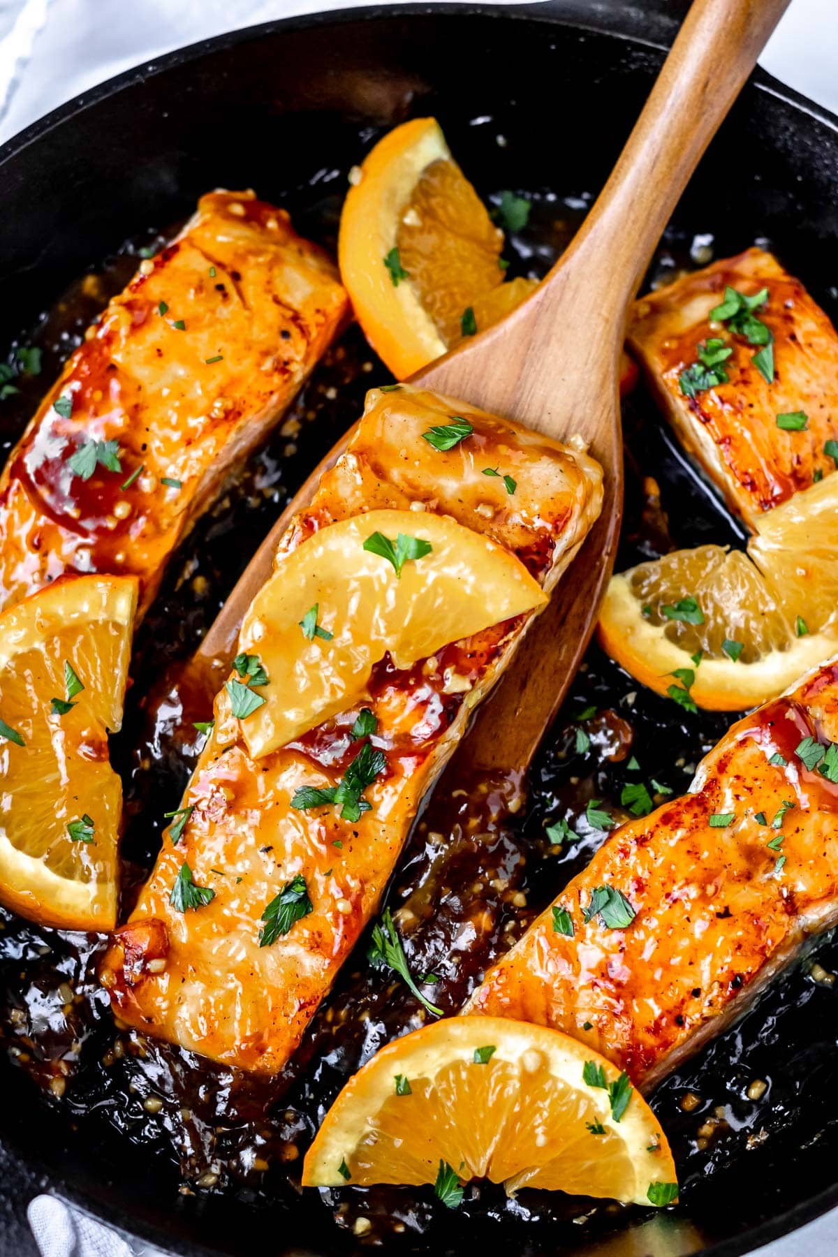 A filet of orange glazed salmon in a cast iron pan being lifted up with a wood turner.