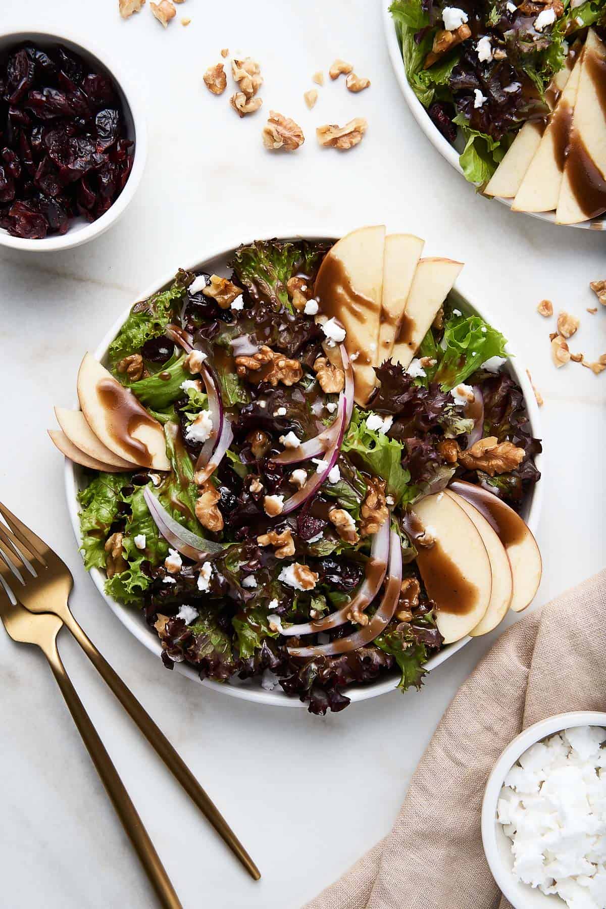 Overhead of an apple walnut salad with a second salad, a bowl of cranberries and gold silverware partially showing around it.