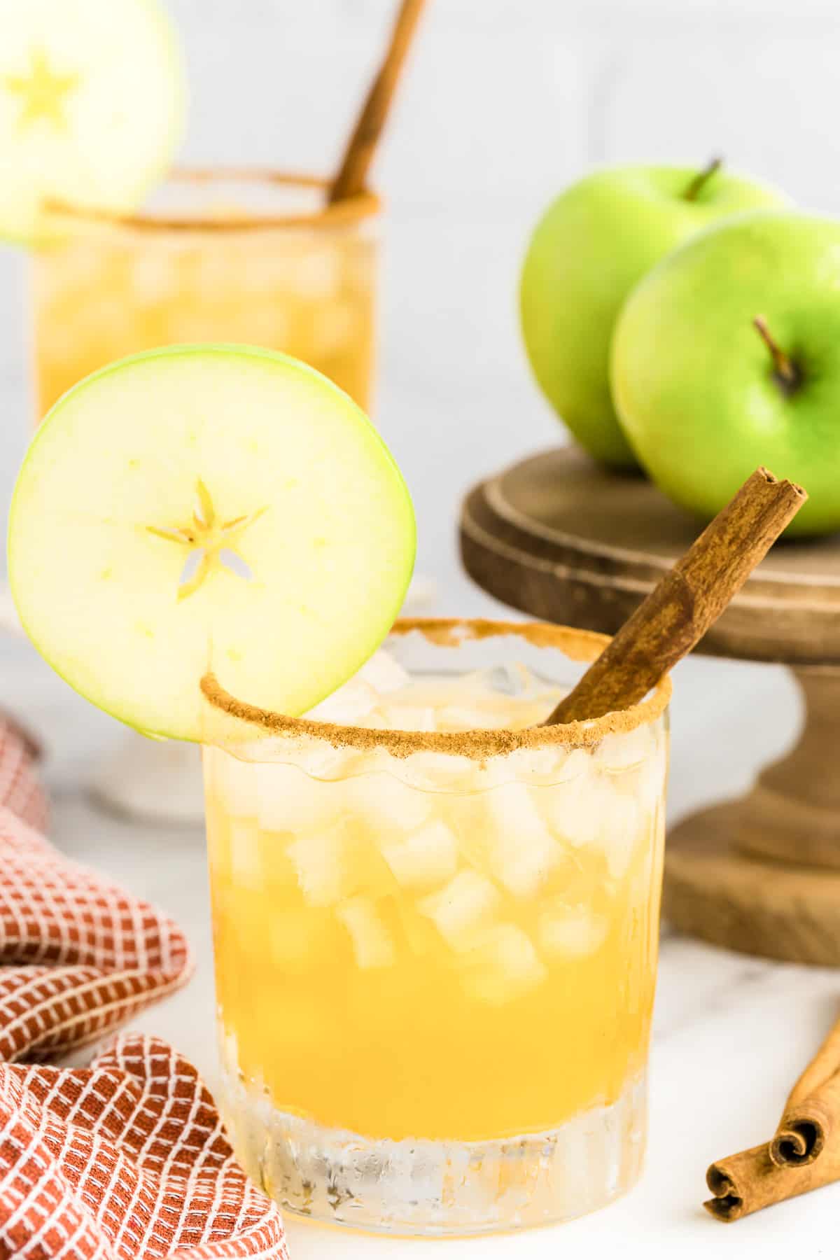 An apple cider margarita with a cinnamon stick in it and a second glass, stand and apples in the background.