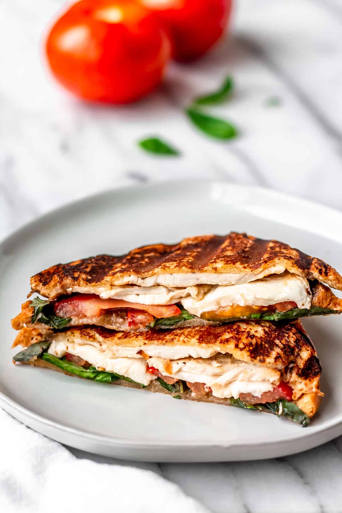 Close up of a Chicken Caprese Panini cut in half and stacked on top of each other on a plate with tomatoes and basil in the background.