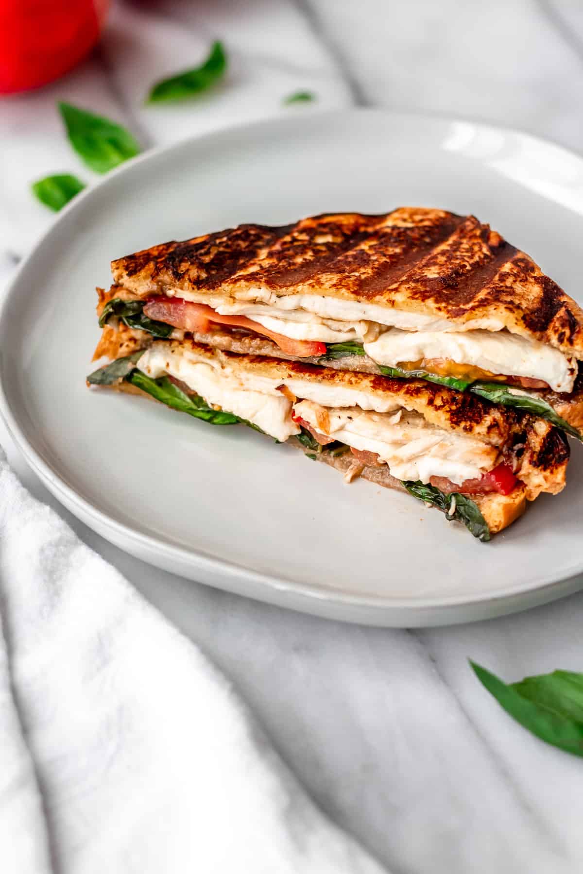 A Chicken Caprese Panini cut in half on a white plate with tomatoes and basil in the background.