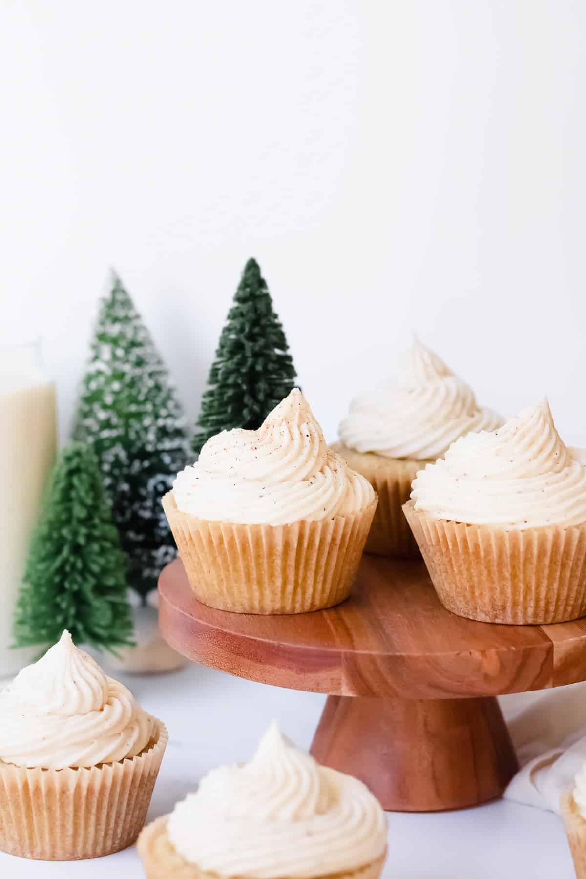 Eggnog cupcakes on a wood stand and in front of it with faux mini christmas trees in the background.