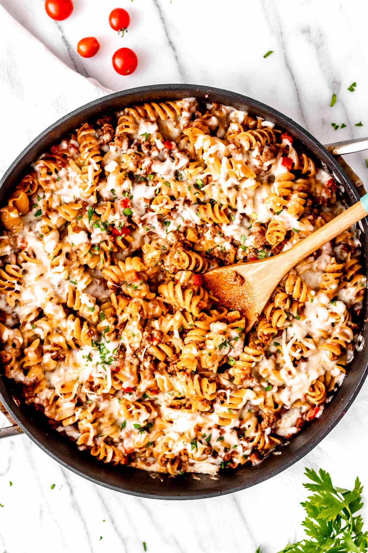 A ground beef pasta skillet on a marble background with tomatoes and parsley around it.