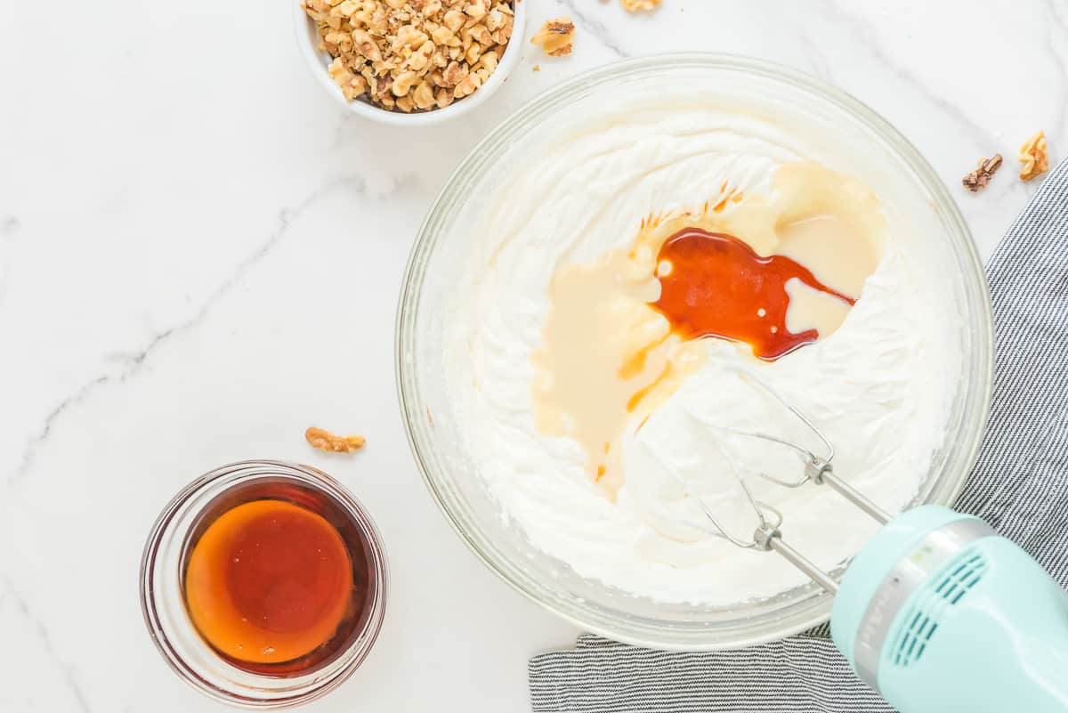 Whipped cream in a mixer bowl with condensed milk and vanilla extra on top with other ingredients around it.