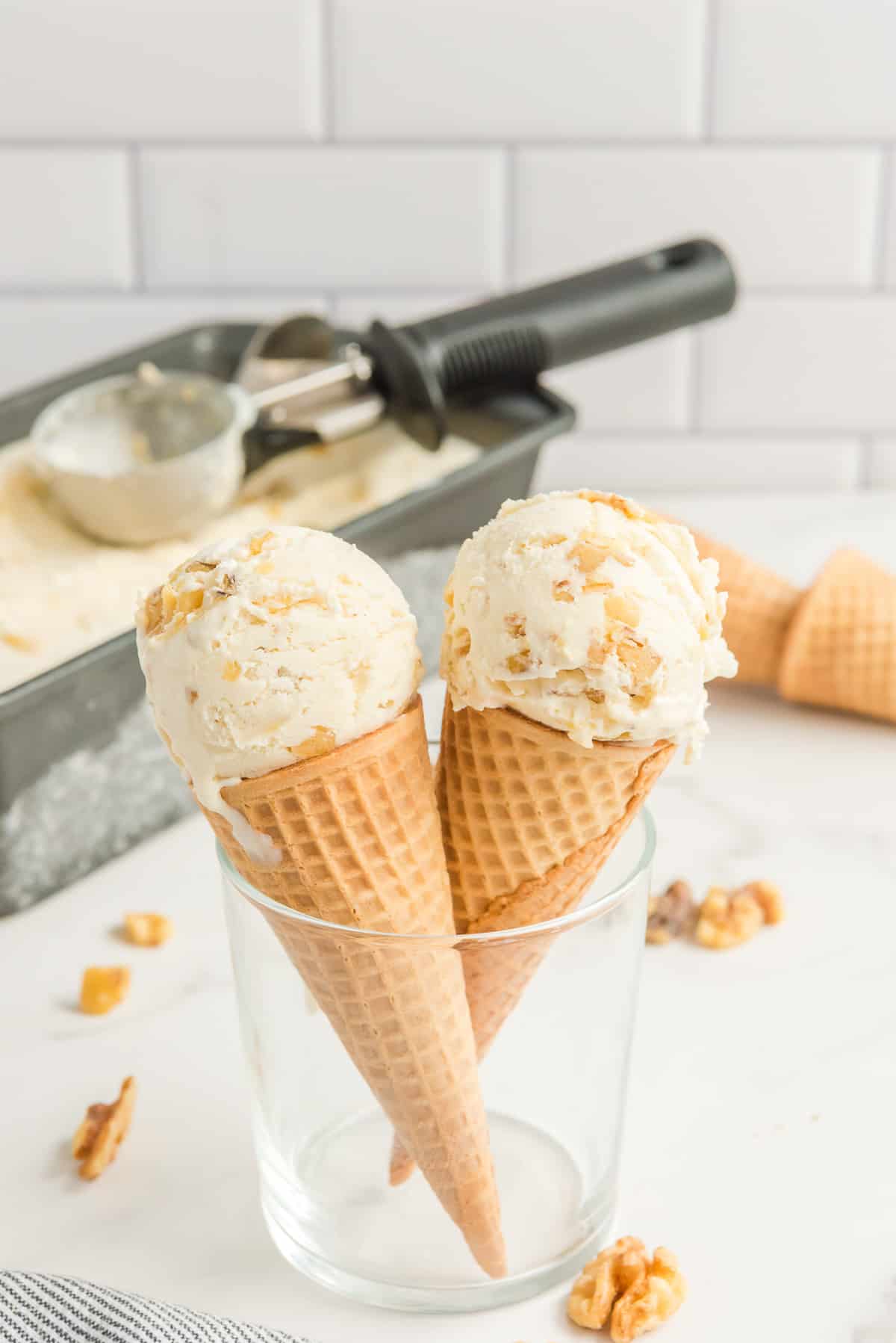 No churn maple walnut ice cream in ice cream cones with a loaf pan filled with ice cream and an ice cream scoop in the background.