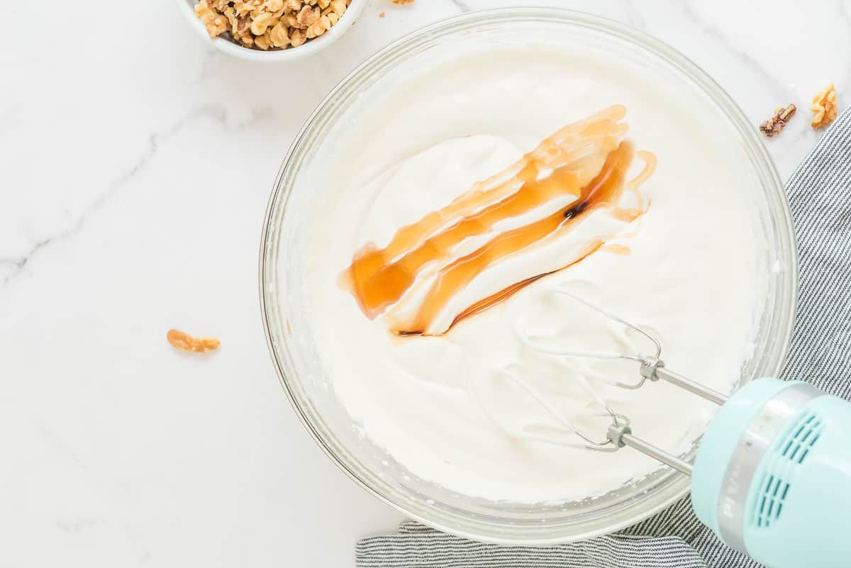 Whipped cream with maple syrup on top in a mixer bowl with a mixer partially showing and walnut around it.