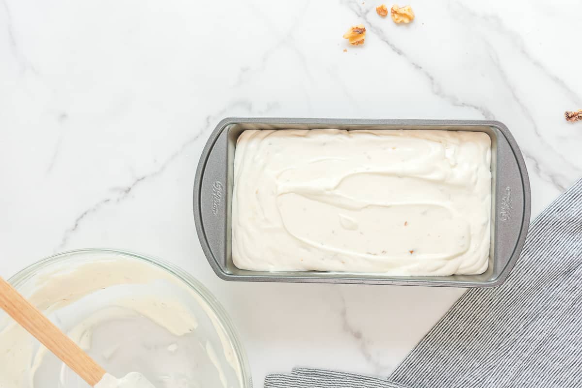 Maple walnut ice cream mixture in a silver loaf pan with an empty bowl and walnuts around it.