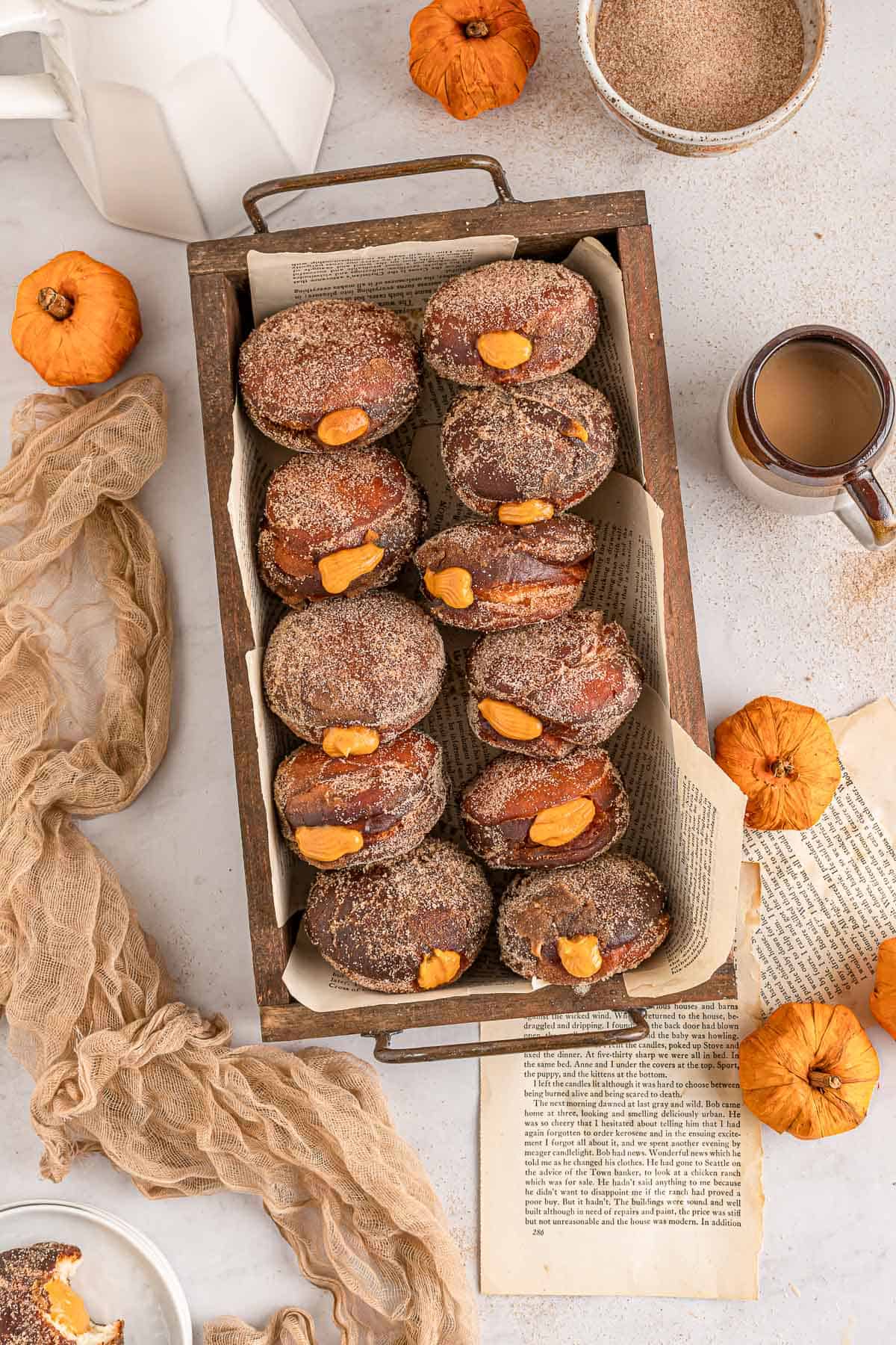 Overhead of pumpkin filled donuts in a wood box with pumpkins, a towel and spices around it.
