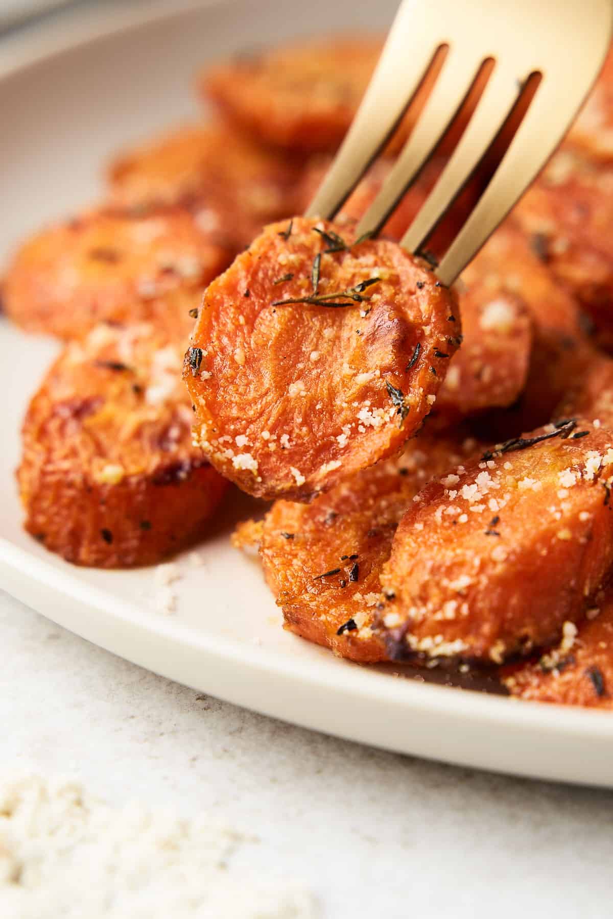 A fork piercing a roasted carrot slice in front of a plate of more carrots.