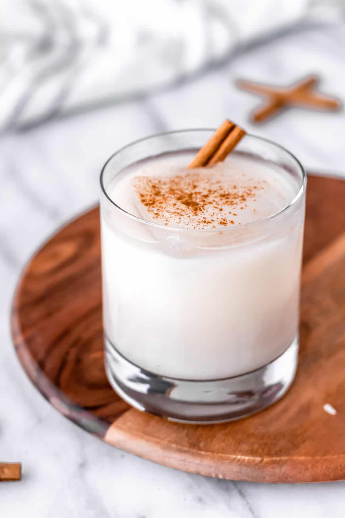 Close up of a glass of horchata with cinnamon sticks and ice in it on a wood server with a striped towel in the background.