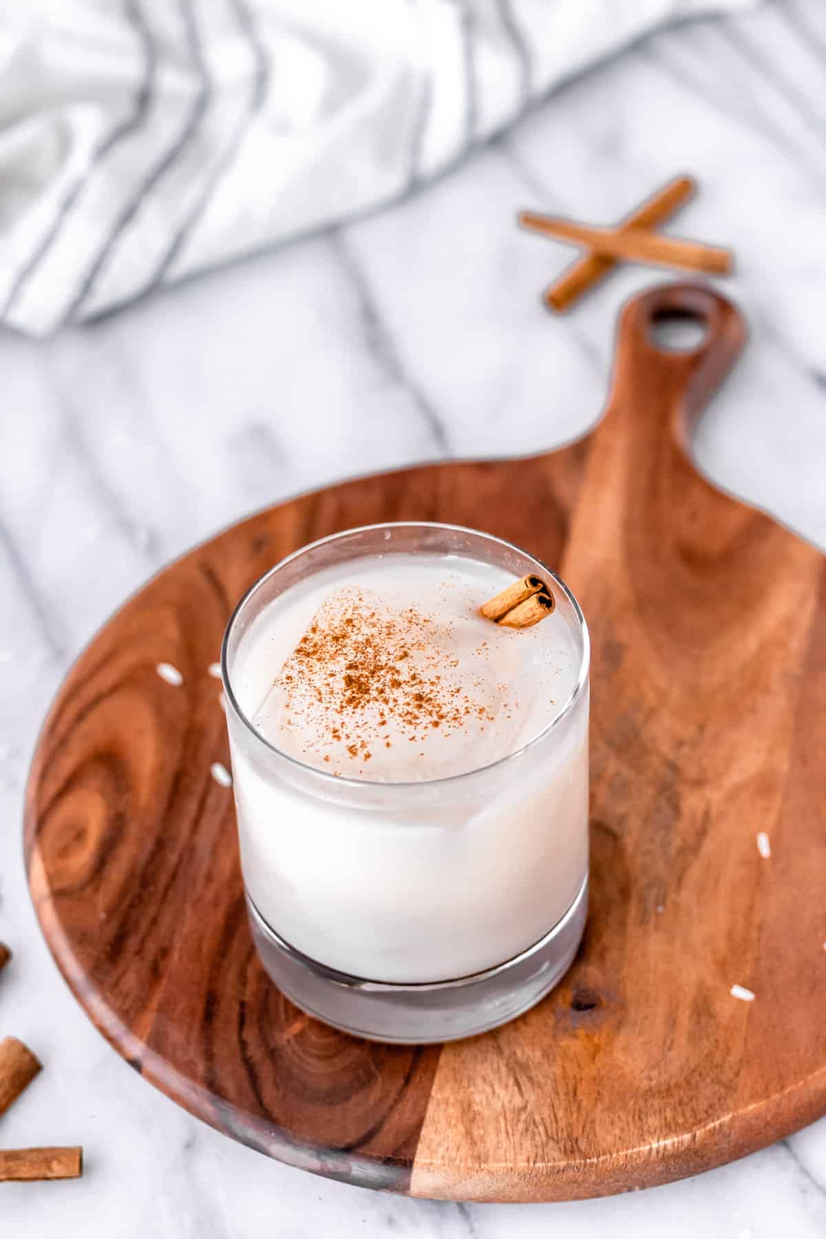 A glass of almond milk horchata on a wood server over a white backdrop with cinnamon sticks and a striped towel in the background.