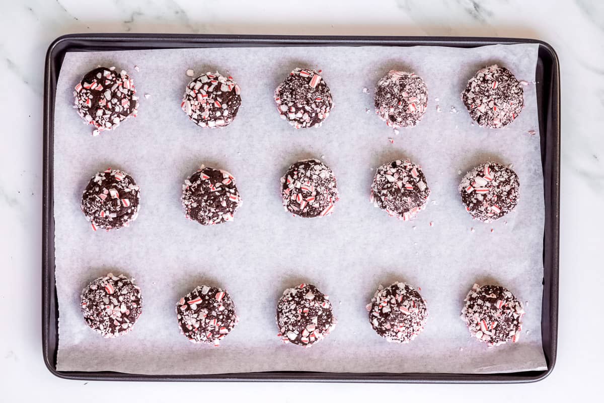Chocolate dough balls coated in crushed candy canes on a parchment paper lined baking sheet.
