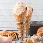 Cinnamon ice cream in cones in metal spiral stands with cinnamon rolls around it and a kitchen towel next to them.