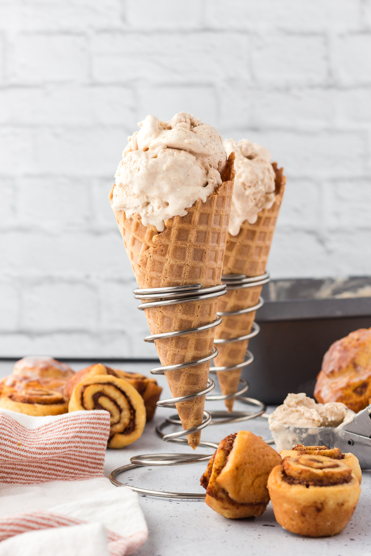 Cinnamon ice cream in cones in metal spiral stands with cinnamon rolls around it and a kitchen towel next to them.
