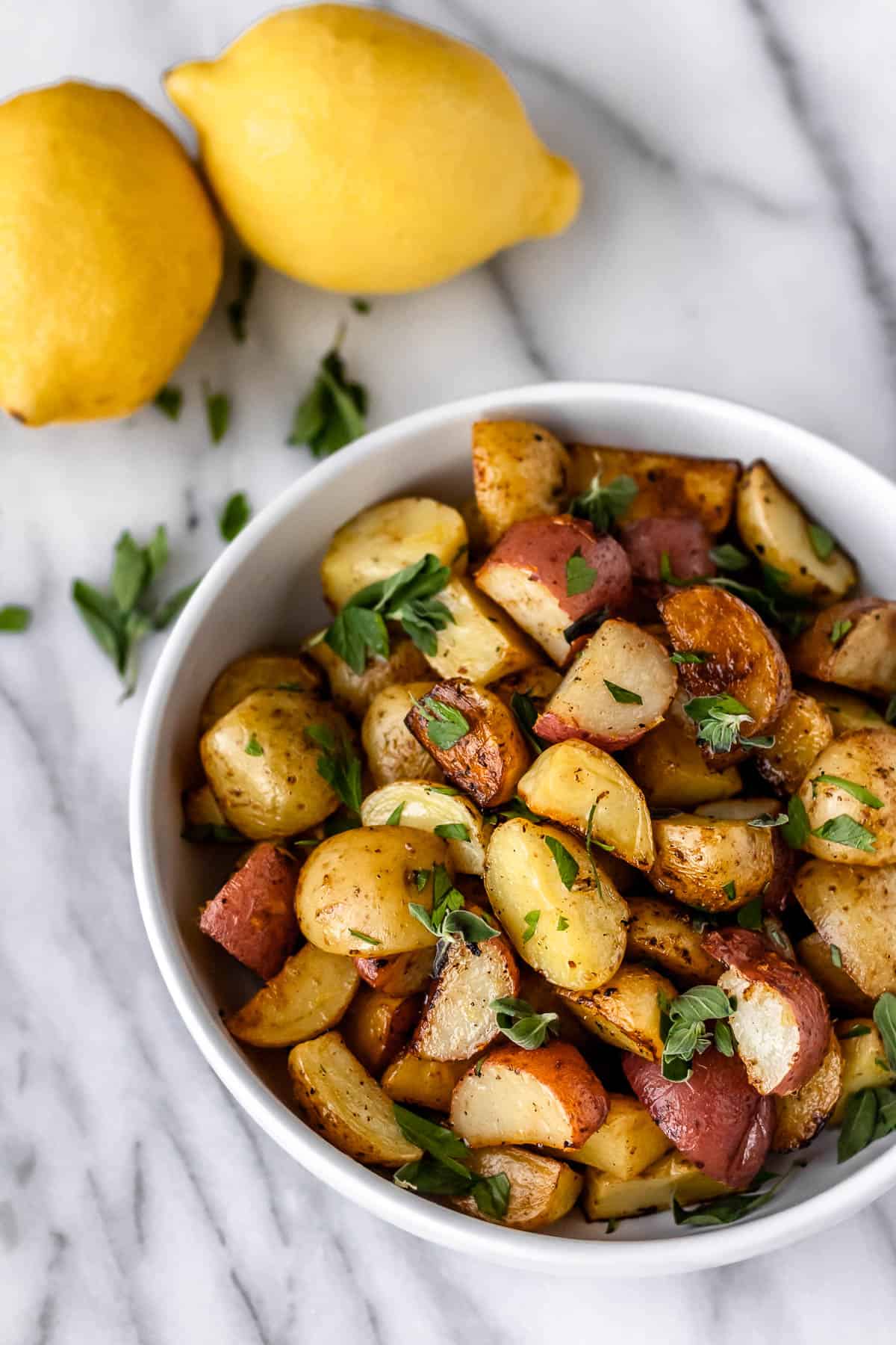A bowl of Greek roasted potatoes with lemons and fresh oregano leaves around it.