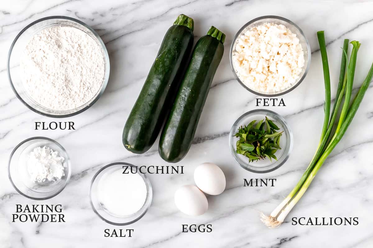 Ingredients needed to make Greek zucchini fritters on a marble background with text overlay.