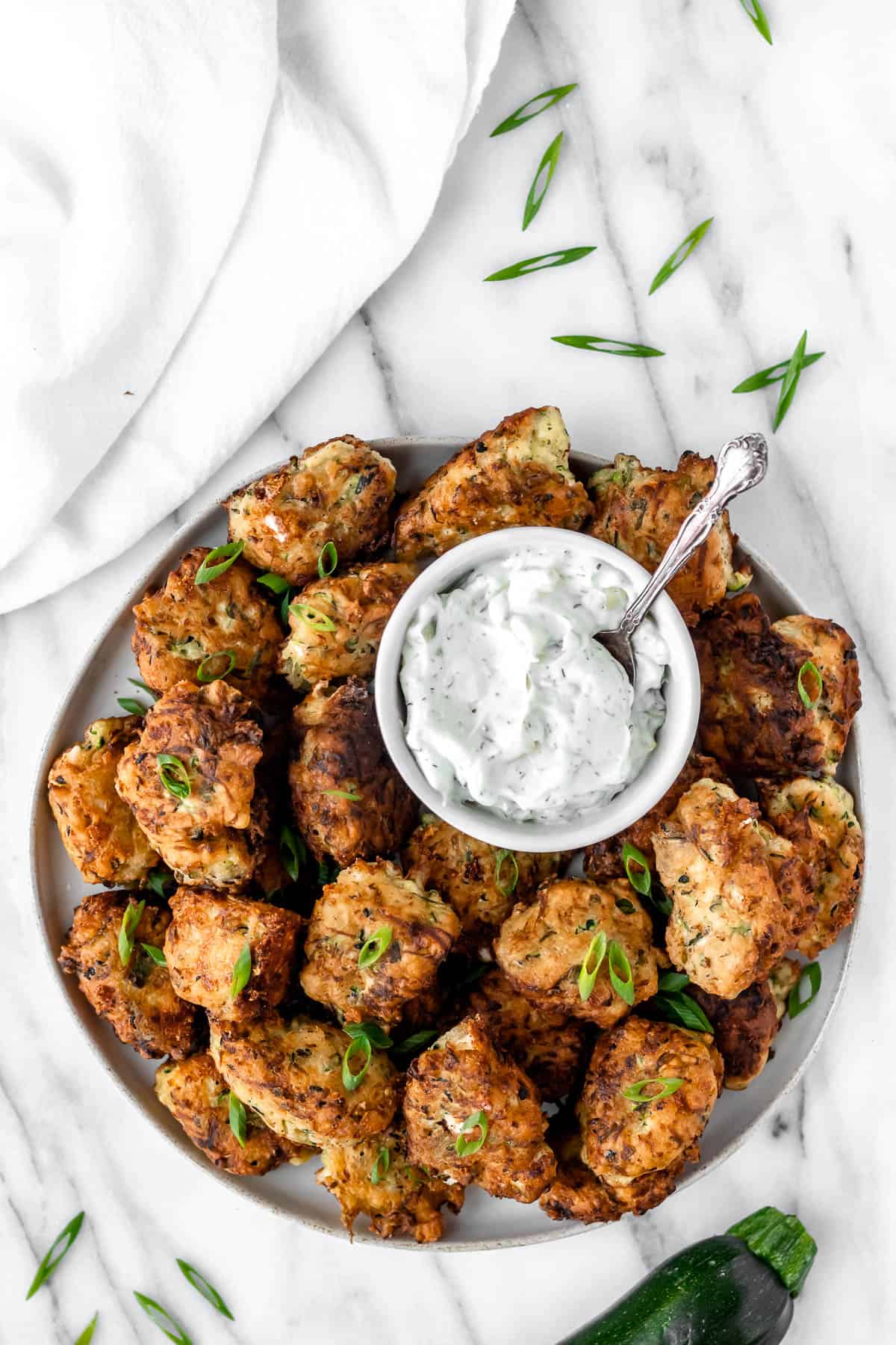 A round plate of kolokithokeftedes with a smaller bowl of tzatziki on it and zucchini and sliced scallions around it.