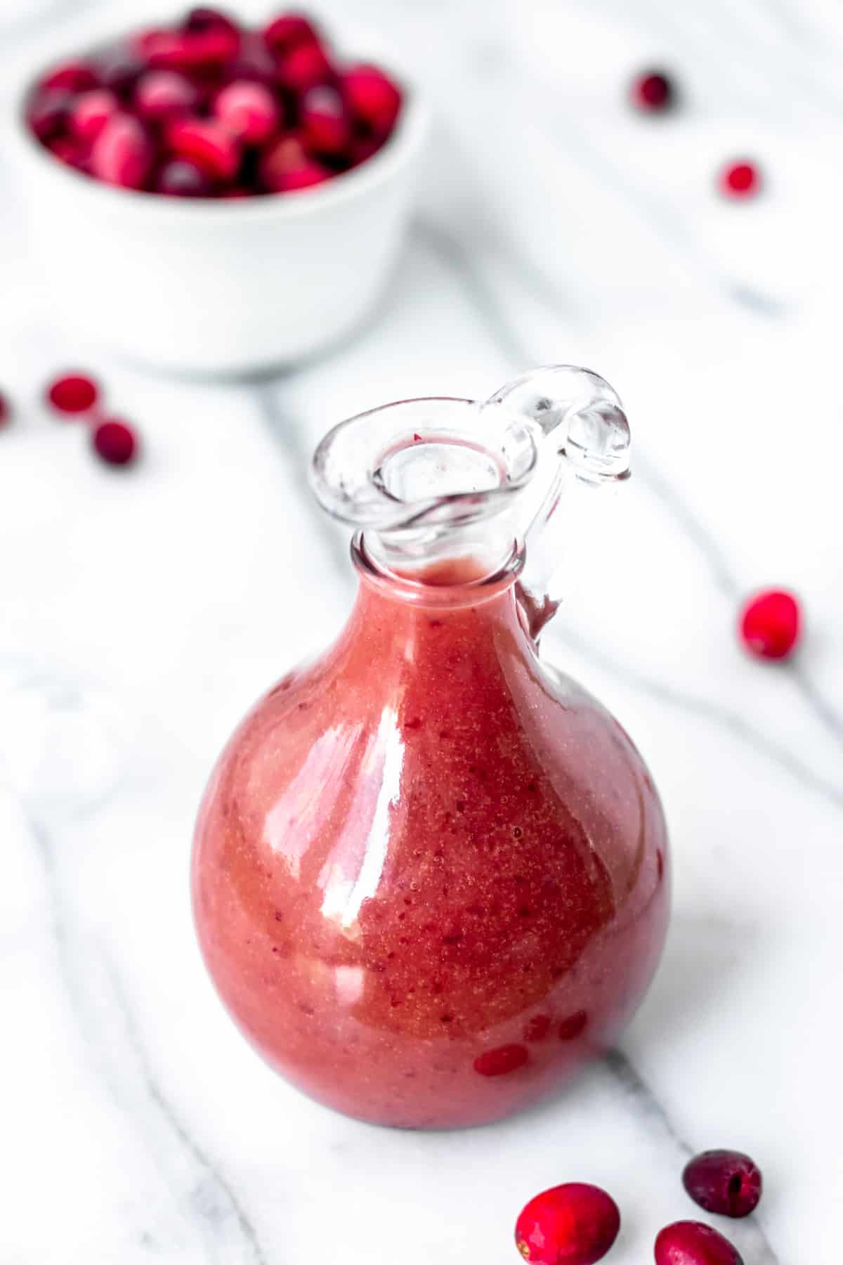 Cranberry salad dressing in a carafe with a bowl of cranberries and loose cranberries around it.