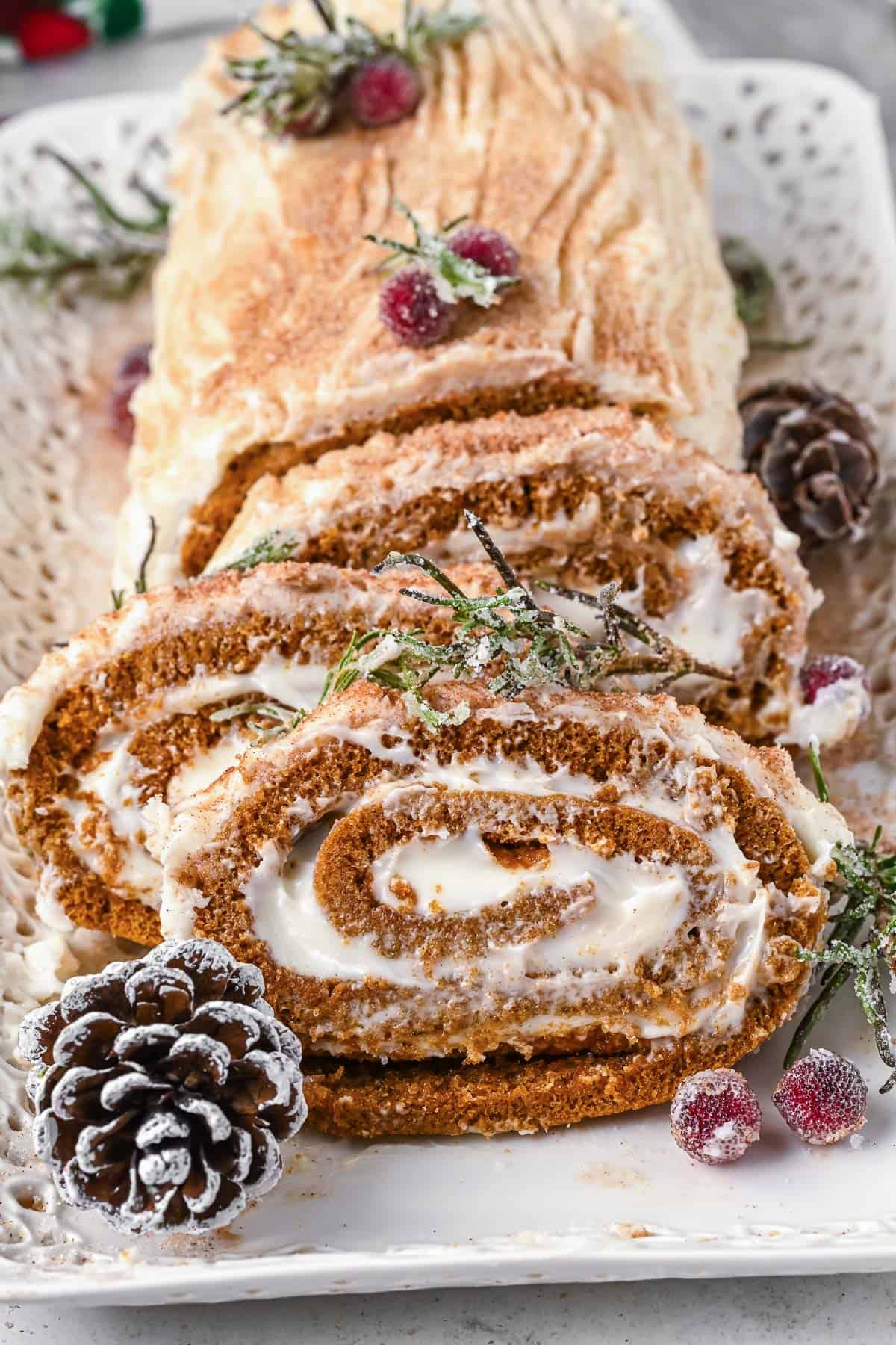 Slices of a gingerbread yule log slanted against the uncut cake with pine cones, sugared cranberries and rosemary for decor.