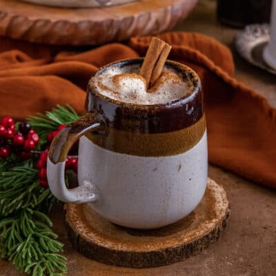 A gingerbread latte in a brown and cream mug with leaves, berries and another mug in the background.