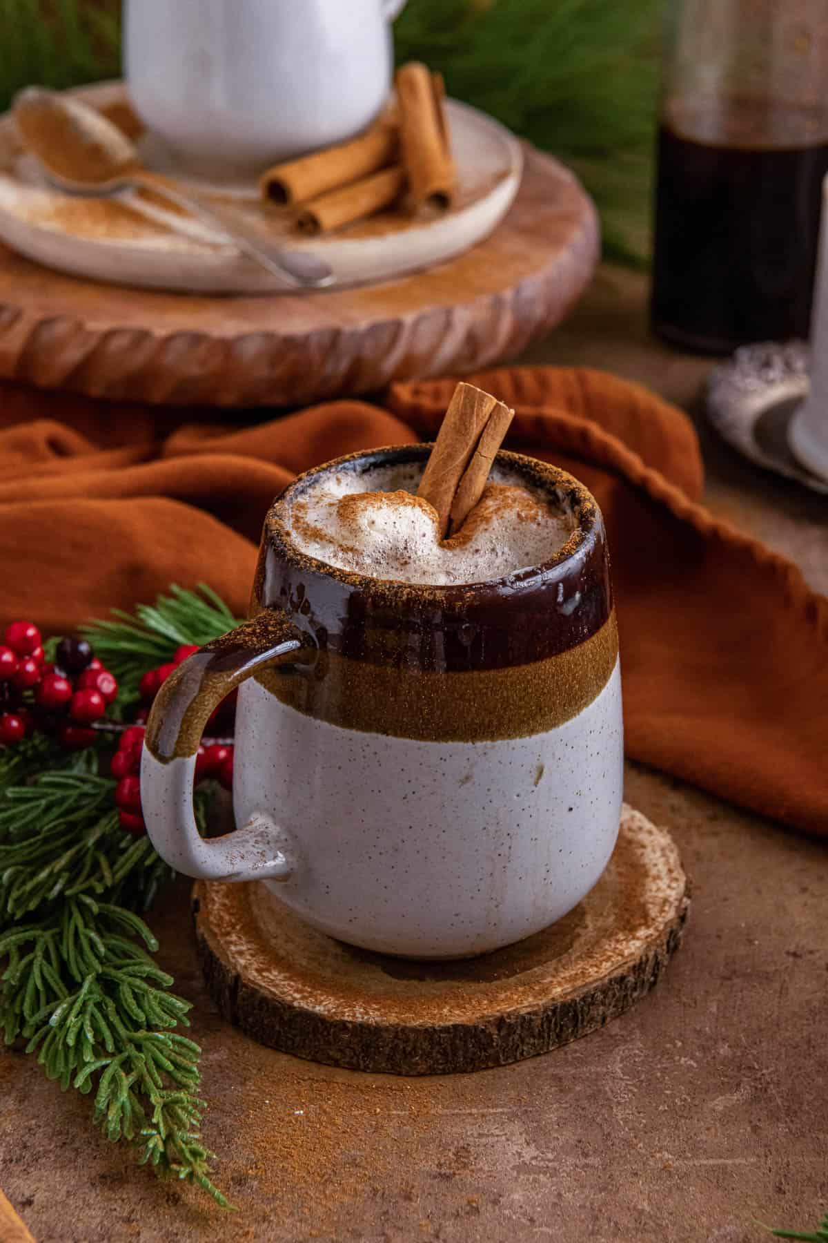 A gingerbread latte in a brown and cream mug with leaves, berries and another mug in the background.