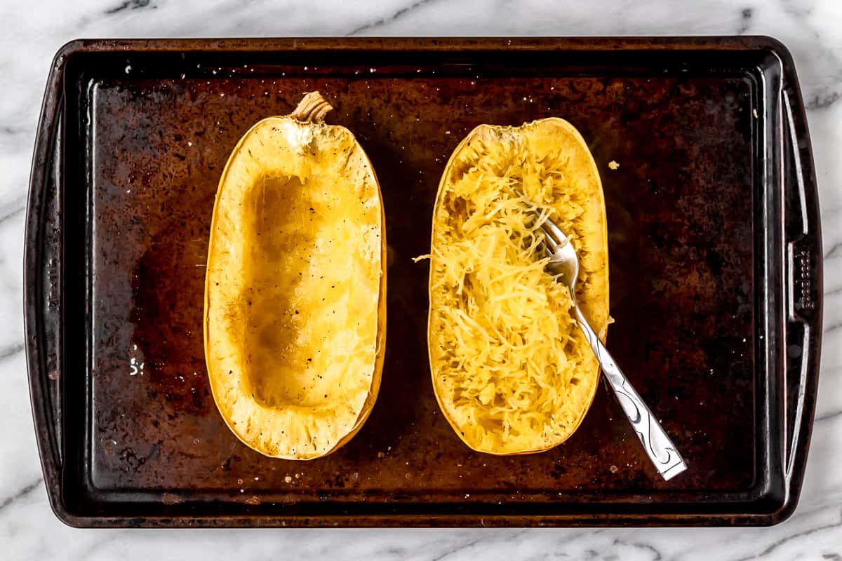 Two halves of roasted spaghetti squash on a sheet pan with the strands scraped from one half with a fork.