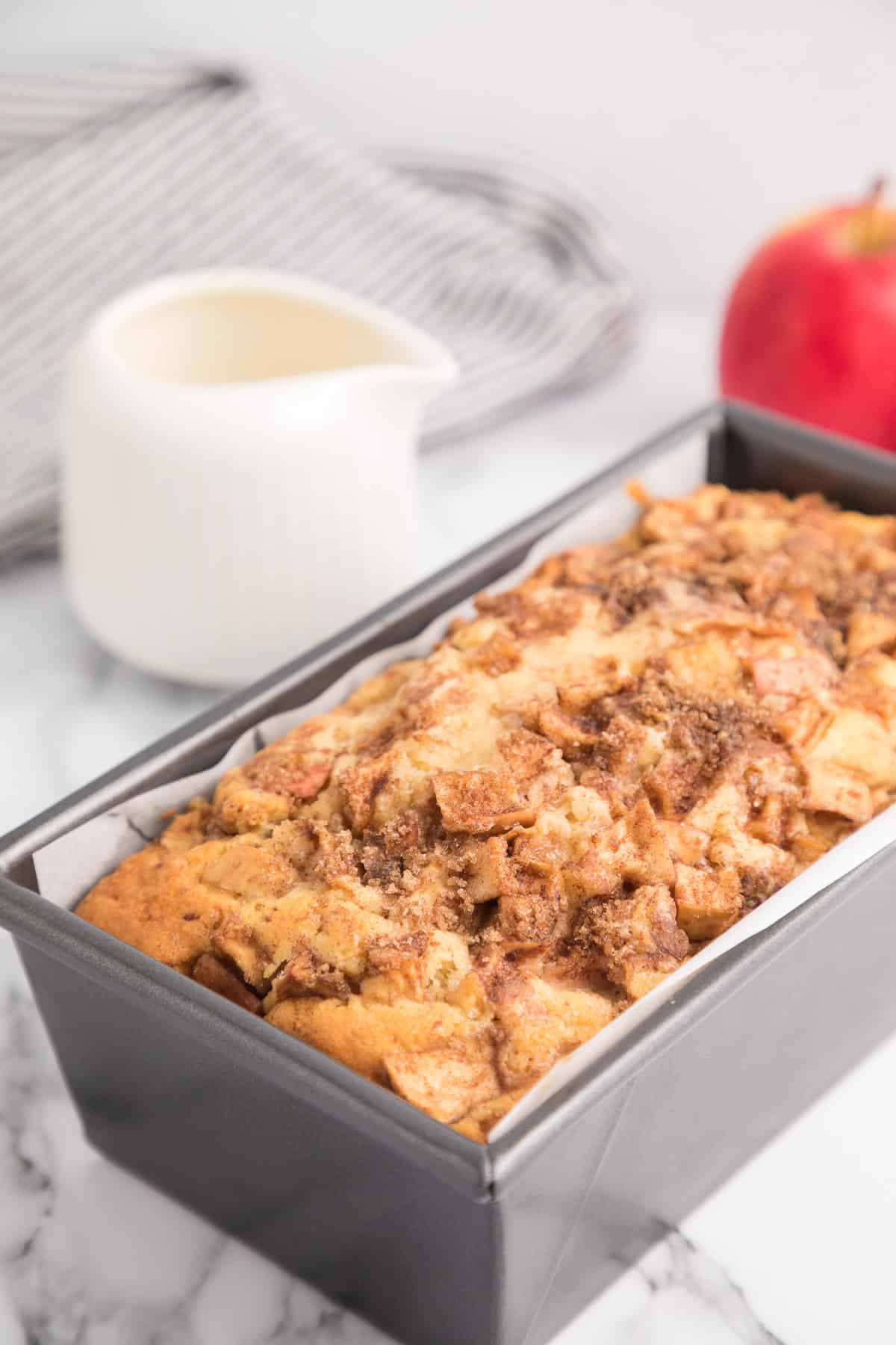 A baked apple fritter bread close up in a pan with a container of glaze and apples in the background.