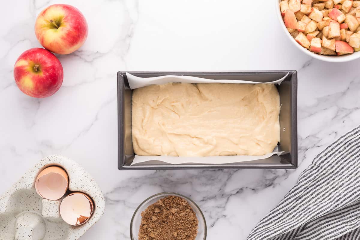 Apple fritter cake batter in a loaf pan with apples and other ingredients around it.