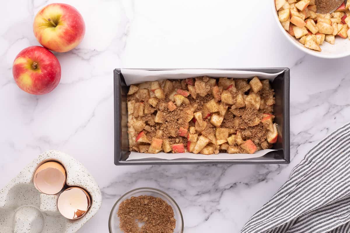 Apple fritter bread batter topped the apples and spices in a loaf pan with apples and other ingredients around it.